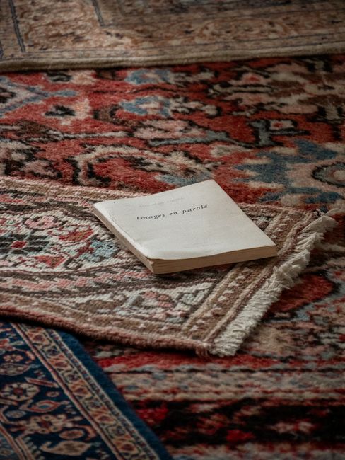 Close-up image of a traditional, patterned area rug with intricate floral and geometric designs in shades of red, blue, and beige, spread across a wooden floor. The rug appears clean, with a slight sh