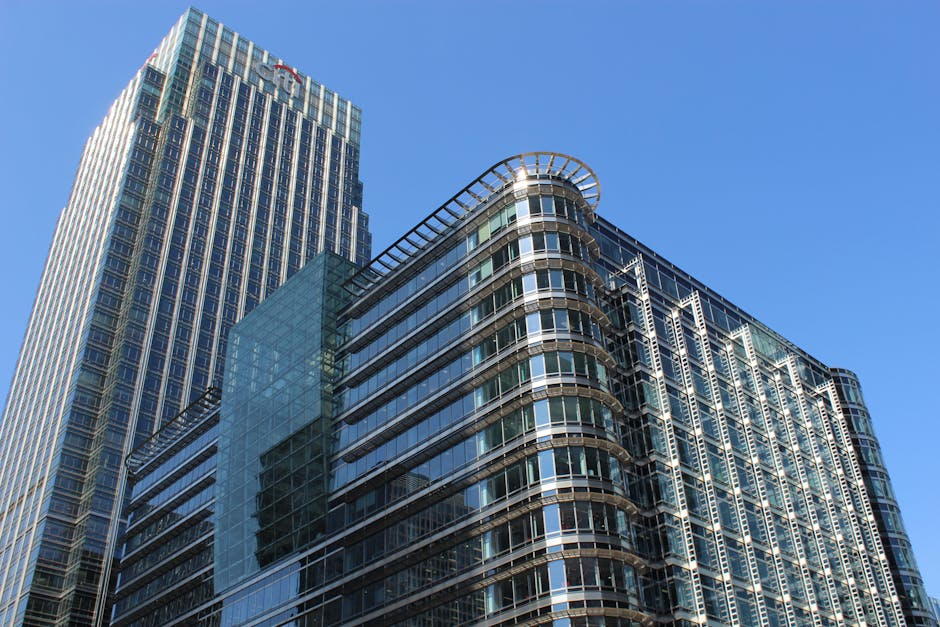Black and white aerial photograph of a modern cityscape highlighting several multi-story office buildings with glass and concrete facades in Canary Wharf. The central building features a distinctive r
